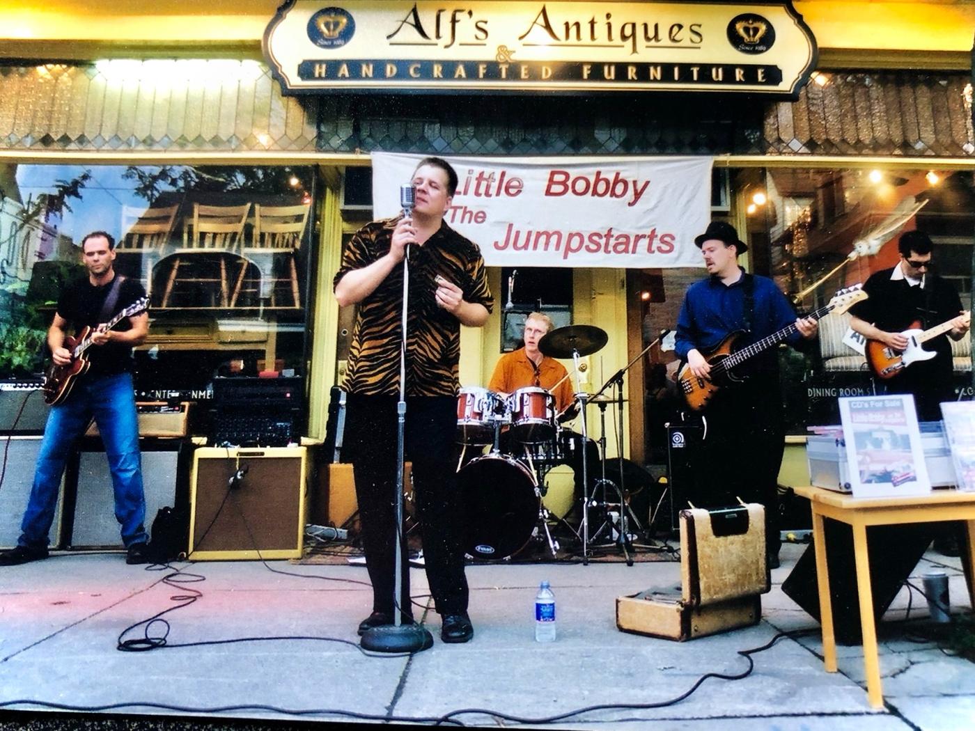 The band playing on a sidewalk in front of an antiques shop, with a Little Bobby and The Jumpstarts banner behind the drummer